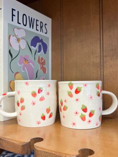 two coffee mugs sitting on top of a wooden table next to a flower book