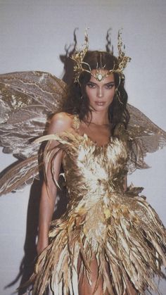 a woman dressed as a fairy with wings on her head and gold dress, standing in front of a white wall