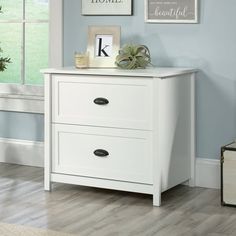 a white dresser with two drawers in front of a window and pictures on the wall