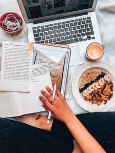 a woman is reading a book next to her laptop and eating food on the bed