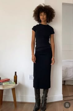 a woman standing in front of a white wall wearing boots and a black dress with short sleeves