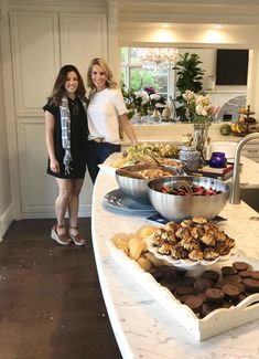 two women standing in front of a table full of food and desserts on it