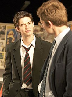 two men in suits talking to each other at an art show or exhibition, with posters on the wall behind them