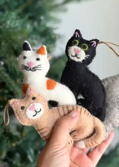 a hand holding three cat ornaments in front of a christmas tree