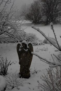 a statue in the snow surrounded by bushes and trees with snow falling on it's branches