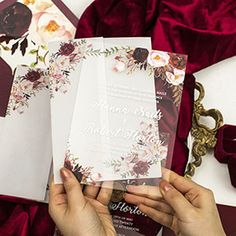 two people holding up wedding cards on top of a red velvet table cloth with flowers