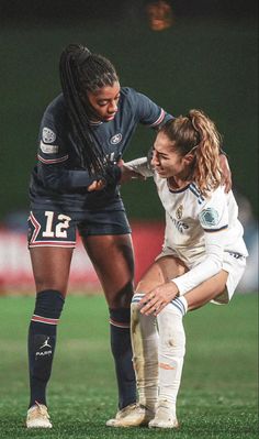 two female soccer players on the field during a game, one is kneeling down and the other has her arm around another player's shoulder