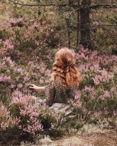 a woman with long red hair sitting in the middle of a flowery field looking at purple flowers