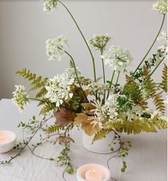 two white candles are sitting on a table with some flowers and greenery in it