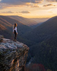 a woman standing on the edge of a cliff looking out over a valley at sunset