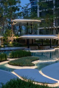 an outdoor swimming pool surrounded by greenery and umbrellas at night with buildings in the background