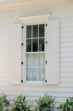 an open window on the side of a white house with green bushes in front of it