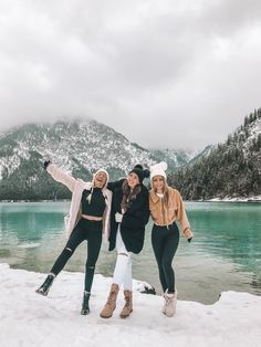 three women posing for the camera in front of a lake with snow covered mountains behind them