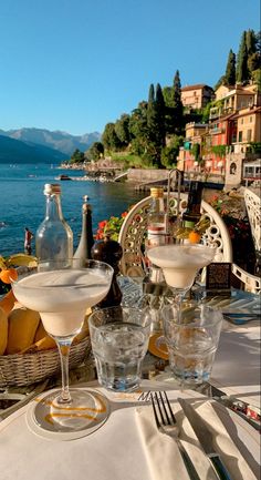 an outdoor dining table with drinks and fruit on it, overlooking the water's edge