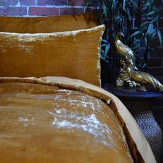 a bed with gold colored sheets and pillows in front of a brick wall next to a plant