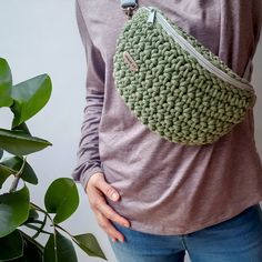 a woman wearing a green crochet purse with her hands in her pockets, standing next to a potted plant