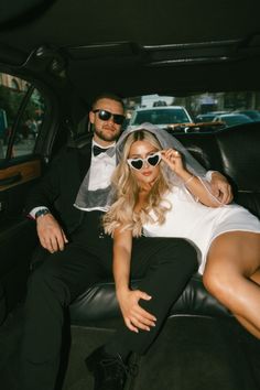 a man and woman sitting in the back of a car wearing black sunglasses, white dress and tuxedo