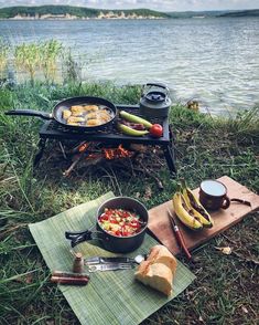 an outdoor grill with food on it next to a lake and picnic mat, near the water's edge