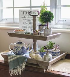 two tiered tray with plants and teapots on it in front of a window