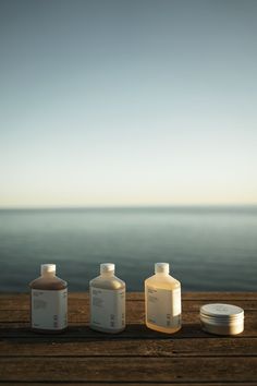 three bottles are lined up on a wooden table near the ocean, one is empty