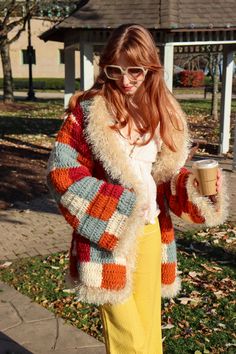 a woman is standing on the sidewalk holding a coffee cup and wearing a multicolored cardigan