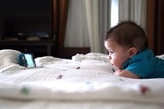 a baby laying on top of a bed next to a cell phone in his hand