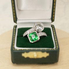 an emerald and diamond ring sits in a velvet box on a wooden table with its lid open