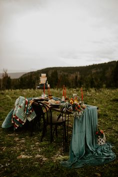 an outdoor table set up with candles, flowers and blankets on the grass in front of a mountain