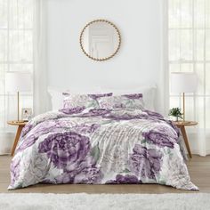 a bed with purple flowers on it in a white room next to two tables and windows