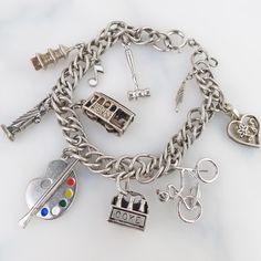 A variety of vintage sterling silver charms dangle from this sterling silver chain bracelet. There are plenty of open links to customize the bracelet with other special acquired charms. This piece weighs in at 31.2 grams and stretches 6 3/4 inches long.  The charms are in varying vintage condition with some vintage patina. My favorite charm on this bracelet is the artist's palette with paintbrush and enameled colors. The other 8 charms included are: a gavel, pagoda, music note, Statue of Liberty, San Francisco Cable car, six pack of Coke, a bicycle, feather and heart with flower. Most are hallmarked as sterling silver; two that weren't tested as sterling silver. Overall, this piece is in very good vintage condition with expected surface age wear to silver and/or patina. There are a few emp Affordable Silver Bracelet With Vintage Charm, Silver Sterling Silver Necklace With Dangling Charms, Vintage Sterling Silver Jewelry With Dangling Charms, Antique Silver Metal Jewelry With Charms, Sterling Silver Dangle Jewelry With Vintage Charm, Sterling Silver Vintage Charm Bracelet As A Gift, Silver Charm Bracelet Aesthetic, Silver Metal Charm Necklaces, Silver Sterling Charm Necklace With Removable Charms