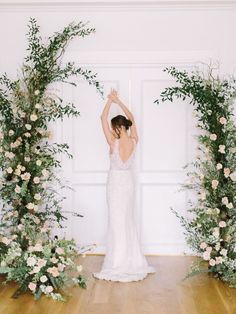 a woman in a wedding dress standing next to flowers and greenery with her arms up