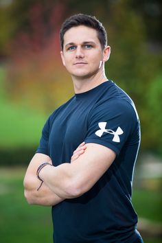 a man with his arms crossed standing in front of some grass and trees, wearing a blue undershirt