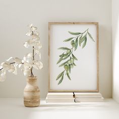 a vase with white flowers sitting next to an art print on a shelf in front of a window