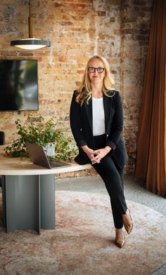 a woman sitting at a table with a laptop in front of her and a brick wall behind her