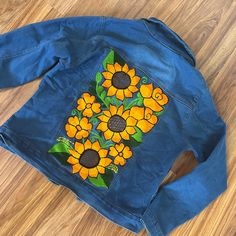 a blue jean jacket with sunflowers painted on the front and back, sitting on a wooden floor