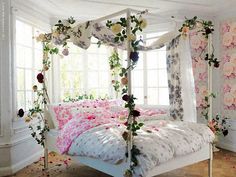 a white bed topped with pink flowers next to a window covered in curtains and drapes