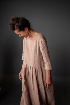 a woman in a beige dress standing against a gray wall with her hands on her hips