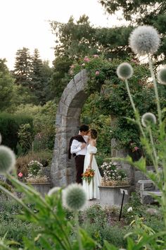 a bride and groom standing in front of a stone arch surrounded by greenery with dandelions