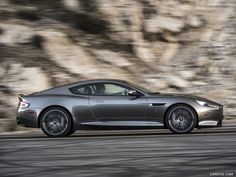 a grey sports car driving down the road in front of a rocky cliff side wall