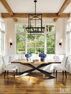 a dining room table with white chairs and a bench in front of two windows that look out onto the woods