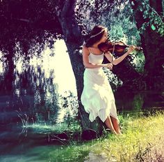 a woman in a white dress is playing the violin by a tree with water behind her