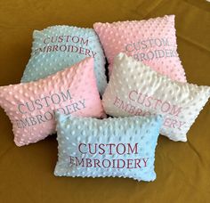 four pillows with custom embroidery on them are sitting on a brown tablecloth, one is pink and the other is blue