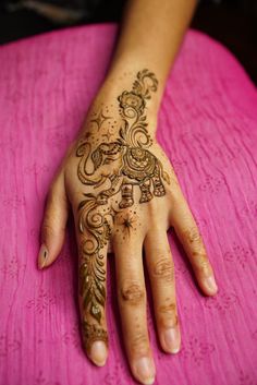 a woman's hand with henna tattoos on her left arm and fingers, sitting on a pink surface