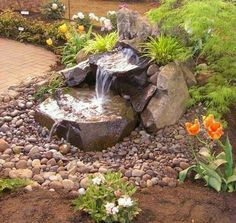 there is a water fountain in the middle of some rocks and flowers on the ground