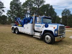 a blue and white tow truck is parked in the middle of a grassy field with trees behind it