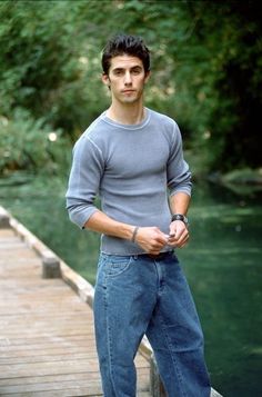 a young man standing on a wooden bridge next to a body of water with trees in the background