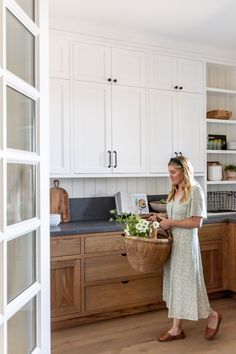 a woman standing in a kitchen holding a basket filled with flowers and greenery on the counter