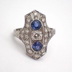 Art Deco 14k (.585) white gold long ring, decorated with Diamonds and Sapphire stones. There are 22 single cut Diamonds, approx. 1.55 mm, the center stone is Old Mine Cut 4.67 x 4.2 x 3.25 mm, color K, SI1 clarity. 2 natural Sapphires are 4.27 mm in diameter. This stately ring is a size 5, it is 22 mm long, weighing 2.7 grams. EA4815 Diamond White Marquise Multi-stone Ring, Diamond White Multi-stone Marquise Rings, White Gold Marquise Multi-stone Diamond Ring, Diamond White Marquise Multi-stone Jewelry, Classic Marquise Multi-stone Diamond Ring, Collectible Sapphire Ring With Center Stone In Platinum, Classic Platinum Gemstone Ring, Diamond White Multi-stone Oval Diamond Ring, Oval Multi-stone Diamond White Diamond Ring