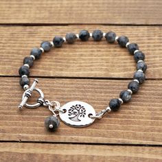 "This bracelet measures 8\" long (20cm) and fits a 6.75\" wrist. It features a silver tone link with a bird in tree design.  I have paired it with black and grey Norwegian moonstone beads (larvikite/black labradorite). The clasp, small beads, and link are made of silver plated leadfree pewter.  The jump rings are made of silver colored enameled copper and antiqued silver plated base metal.  The crimp beads, wire guardian, and crimp covers are made of sterling silver.  The headpin is made of antiqued silver plated base metal.  The bracelet is strung on Soft-Flex wire which is supple and strong. The tree component is made in the US.  It measures 3/4\" long (19mm). ---------------------------- Thank you for browsing my shop! If you'd like to see my necklaces and jewelry sets, click here: http Gray Jewelry With Silver Beads As A Gift, Gray Jewelry With Silver Beads For A Gift, Gray Silver Beaded Jewelry For Gifts, Gray Jewelry With Silver Beads For Gifts, Adjustable Gray Metal Jewelry, Adjustable Gray Sterling Silver Jewelry, Silver Charm Bracelet With Gemstone Beads, Adjustable Silver Charm Bracelet With Gemstone Beads, Adjustable Silver Rosary Bracelet With Gemstone Beads