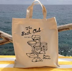 a tote bag sitting on top of a yellow towel next to the ocean with wine glasses